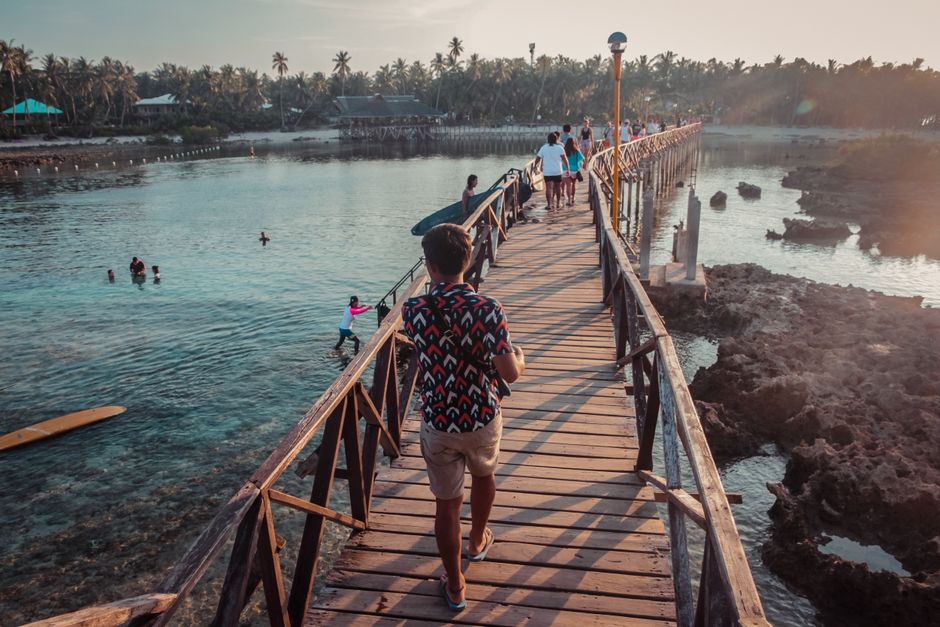 People walk across a scenic wooden bridge in General Luna, Philippines at sunset. (photo by John Matthew on Pexels)