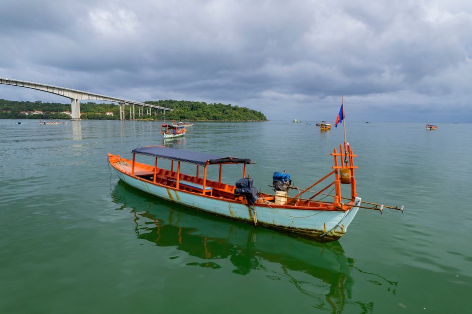 A traditional boat floats in Sihanoukville Bay with a bridge in the background. (photo by Nara Tsitra on Pexels)